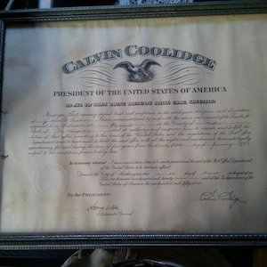 May include: Framed document from the President of the United States, Calvin Coolidge. The document is in black ink on aged paper, with an ornate frame. The text includes the presidential seal and signature.