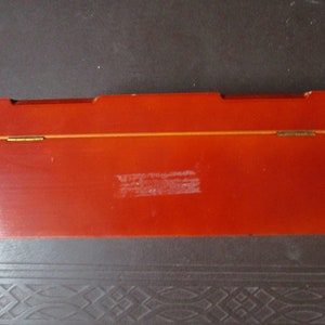 Pu&ograve; includere: Una scatola rettangolare in legno, di colore rosso-arancio, con coperchio incernierato. La scatola ha una finitura lucida e una sottile linea dorata lungo il bordo superiore. Le cerniere sono color ottone. La scatola sembra vuota.