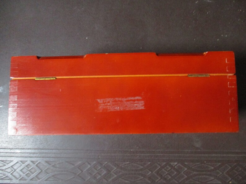 Pu&ograve; includere: Una scatola rettangolare in legno, di colore rosso-arancio, con coperchio incernierato. La scatola ha una finitura lucida e una sottile linea dorata lungo il bordo superiore. Le cerniere sono color ottone. La scatola sembra vuota.