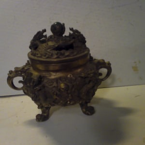 May include: A gold-colored incense burner with intricate dragon designs and a green stone knob on the lid. The burner has three legs and is decorated with floral patterns.