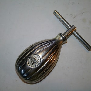 May include: A vintage silver-toned metal tool with a ribbed, bulbous handle and a T-shaped bar. The handle has a circular emblem with the letters "HMO". The tool is likely used for a specific mechanical purpose.