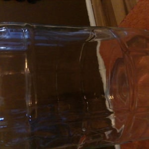 May include: A large, clear glass container with a rounded, barrel-like shape. The glass has a slight greenish tint and is reflective, with visible vertical and horizontal lines. The container is empty and sits on a brown surface.