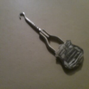 May include: A silver metal shoe horn with a hook at one end and a decorative design at the other end.