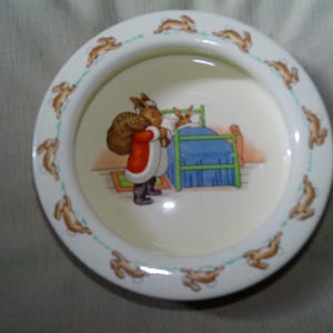 May include: A white ceramic bowl with a rim decorated with brown rabbits. The center features a colorful illustration of a rabbit in a red coat and a sack, visiting another rabbit in bed. A green line connects the rabbits.