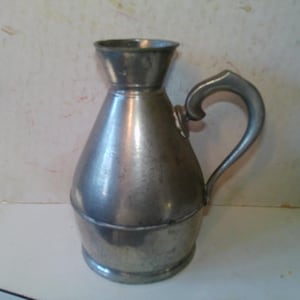 May include: A vintage metal pitcher with a handle. The pitcher has a wide base that tapers to a narrower neck and flared rim. The handle is curved and attached to the side of the pitcher. The metal has a dull, silver-gray color.
