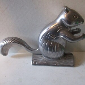 May include: A silver-colored metal squirrel nutcracker. The squirrel is in a sitting position, with its tail curved upwards. The nutcracker is mounted on a base with a wood-like design.