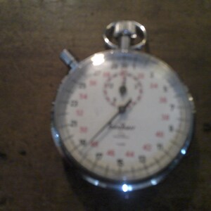 May include: A silver stopwatch with a white face and black hands. The face has red markings for the seconds and minutes. The stopwatch has a black button on the top.