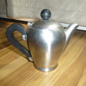 May include: A stainless steel teapot with a black handle and lid knob. The teapot has a curved handle and a spout for pouring. The lid is hinged and has a small black knob. The teapot is sitting on a wooden surface.