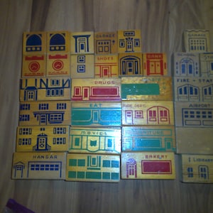 May include: A set of wooden blocks with different building designs painted on them. The blocks feature various buildings, including a garage, school, store, fire station, airport, hangar, bakery, library, and more. Each block has a different color scheme and design.