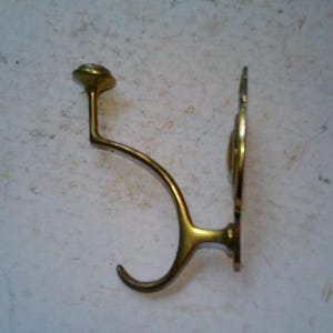 May include: A vintage brass wall hook with a curved arm and decorative backplate. The hook has a rounded end and a small, circular top. The brass has a warm, golden hue, suggesting an antique or retro style. The hook is designed for hanging items.