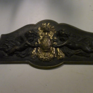 May include: A black metal plaque with two cherubs flanking a gold-painted vase with floral details. The plaque has a scalloped edge and a raised design.