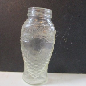May include: Clear glass jar with a textured, fish-scale design. The jar has a wide mouth and a rounded body, tapering towards the base. The jar is empty and sits on a white surface against a dark background.