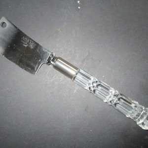 May include: A silver cheese knife with a clear crystal handle. The handle has a faceted design.