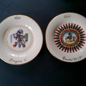 May include: Two small, white decorative plates with gold rims. One plate features a colorful illustration of a figure in traditional garb, the other has a central design with text and a red and white border. The plates have text at the bottom.