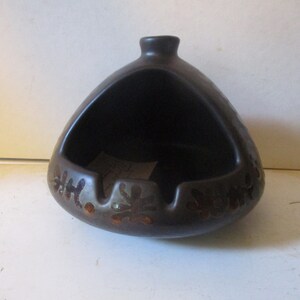 May include: A brown ceramic ashtray with a unique design. The ashtray has a wide, open top and a small, round base. The design features a pattern of stylized leaves and flowers in a lighter brown color.