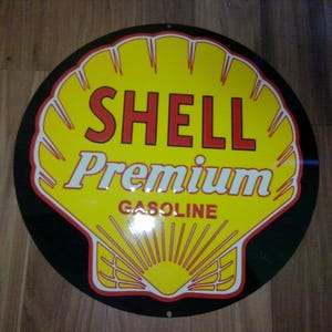 May include: A round Shell Premium Gasoline sign. The sign features a yellow shell shape with red and white text that reads "SHELL Premium GASOLINE" against a black background. The shell design has red and white trim.