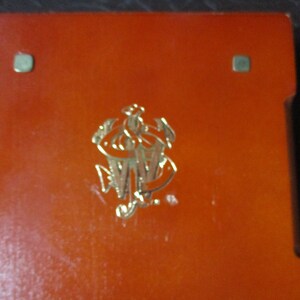Pu&ograve; includere: Una scatola rettangolare arancione con un emblema dorato al centro. L'emblema sembra essere un monogramma o uno stemma stilizzato. Sono visibili due piccoli accenti metallici quadrati.