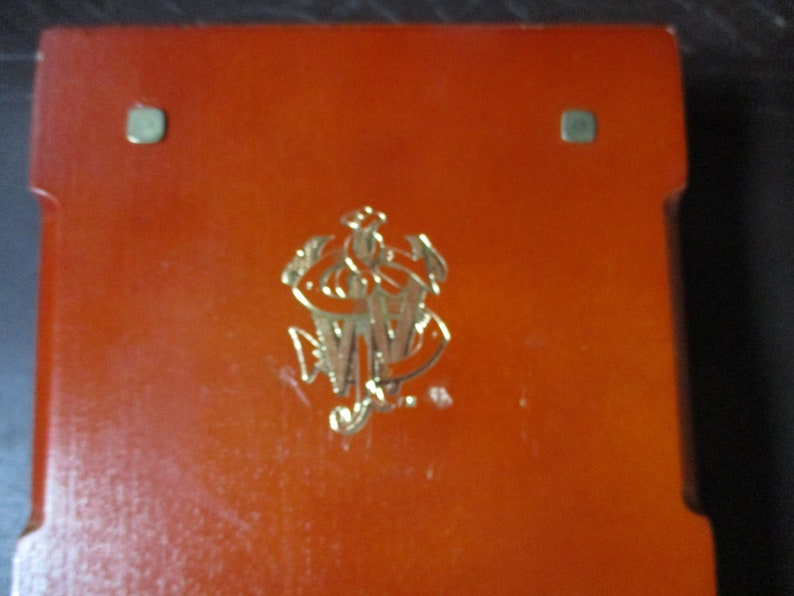 Pu&ograve; includere: Una scatola rettangolare arancione con un emblema dorato al centro. L'emblema sembra essere un monogramma o uno stemma stilizzato. Sono visibili due piccoli accenti metallici quadrati.