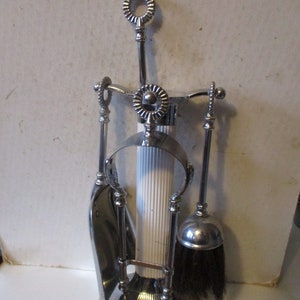 May include: A silver fireplace tool set with a shovel, brush, and poker. The set has decorative handles and a circular base. The brush has black bristles. The set is designed to hold the tools upright.