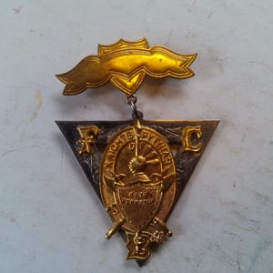 May include: Vintage Knights of Pythias fraternal order pin. The pin features a gold banner, a silver and gold triangle with the letters FPC, and a central gold emblem with a knight's helmet and crossed swords. The pin is likely made of metal.