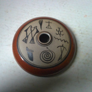 May include: A round, brown ceramic inkwell with a hole in the center. The inkwell is decorated with black and white symbols, including a footprint, a bird, a spiral, and a cross.