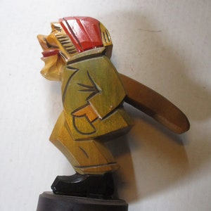 May include: A wooden nutcracker figurine with a red hat, green jacket, and brown pants. The nutcracker is holding a wooden nutcracker in its right hand. The figurine is standing on a round wooden base.