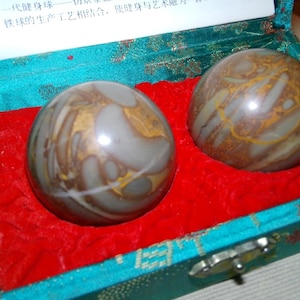 May include: Two patterned stone exercise balls in a teal box with a red velvet lining. The balls have a mix of gray, brown, and gold tones. The box has a silver clasp and a white paper with Chinese text.