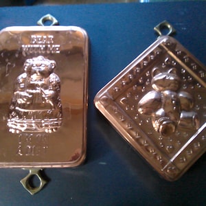 May include: Two antique copper cake molds. One mold features a woman in a dress with the text "Dear Mother" and "Love & Joy". The other mold features a teddy bear.