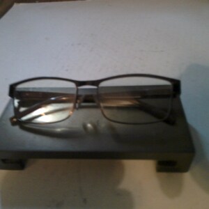 May include: A pair of brown eyeglasses with a metal frame and clear lenses. The glasses are resting on a black display stand.