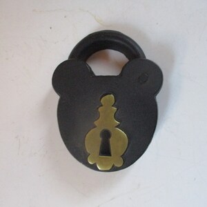 May include: A black padlock with a rounded top and a brass-colored keyhole cover. The padlock has a unique shape with rounded edges, resembling a bear's head. The keyhole cover has an ornate design.
