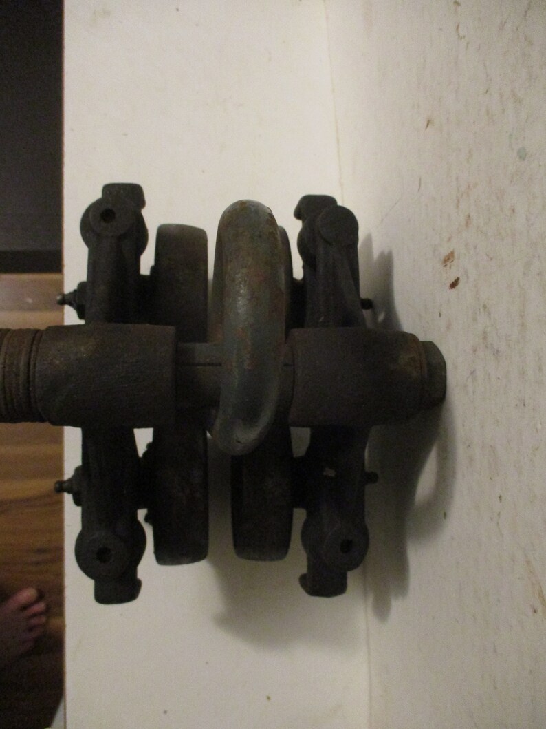 May include: A close-up of a dark, metallic mechanical component. The object has a series of cylindrical and curved parts, with a central ring-shaped element. The metal shows signs of age and wear, with a slightly rusted appearance. The component is mounted against a white wall.