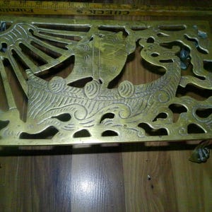 May include: A rectangular brass trivet featuring an openwork design of a sailing ship on waves with a sunburst in the corner. The trivet stands on four small feet and is positioned next to a yellow ruler measuring approximately 12 inches in length.