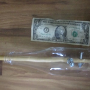 May include: A clear glass bottle with a wooden dowel and metal hardware inside, next to a one-dollar bill. The bottle is lying on a wooden surface.