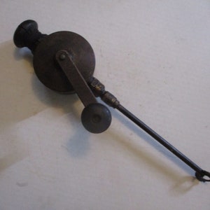 May include: A vintage hand-cranked grease gun with a black handle and a long metal tube. The gun has a round metal body with a crank handle and a small metal nozzle at the end of the tube.