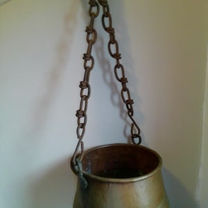 May include: A vintage copper cauldron with a rounded base and a wide opening, suspended by a chain. The cauldron has a tarnished, aged appearance, suggesting it is antique. The chain is made of dark metal links.