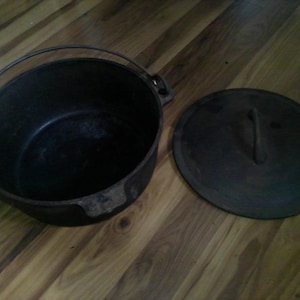May include: A black cast iron Dutch oven with a lid and handle. The pot has a handle for carrying and the lid has a handle for lifting. The pot and lid are sitting on a wooden surface.