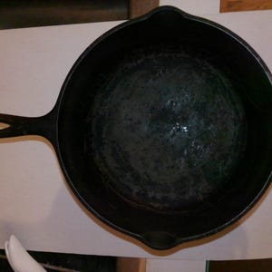 May include: A black cast iron skillet with a long handle and a rounded cooking surface. The skillet has a well-seasoned interior and is designed for stovetop or oven use. The skillet is shown from a top-down perspective.