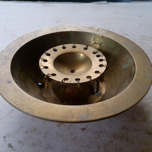 May include: A vintage brass bowl with a central, circular insert. The insert has a series of small holes around its perimeter, connected by small chains. The bowl has a wide, shallow design, and the brass has a warm, aged patina.
