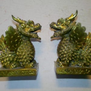 May include: A pair of gold-colored dragon statues with intricate details. Each dragon is perched on a square base with decorative carvings. The dragons have outstretched wings and are facing opposite directions.