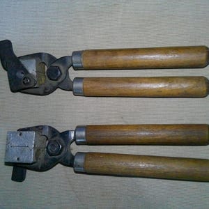 May include: Two metal bullet mold tools with wooden handles. Each tool features a dark metal hinge mechanism connected to two cylindrical wooden grips. The metal heads have a square block shape with a small horizontal slit visible on the face.