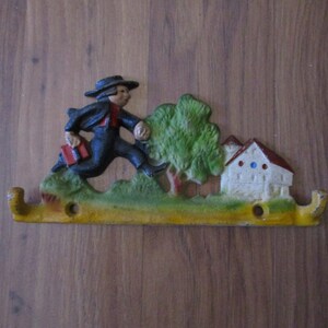May include: A cast iron wall hook with a yellow base and green and brown accents. The hook features a figure of a person running with a book in hand, a small house, and a tree.