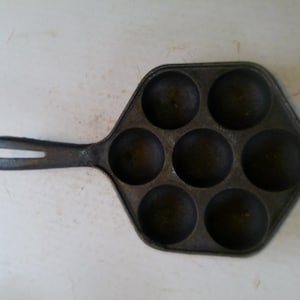 Może przedstawiać: Żeliwna patelnia w kolorze ciemnoszarym z siedmioma okrągłymi wgłębieniami do gotowania. Patelnia ma długi uchwyt z otworem do zawieszania. Powierzchnia patelni sugeruje, że jest dobrze wypalona i gotowa do użycia.