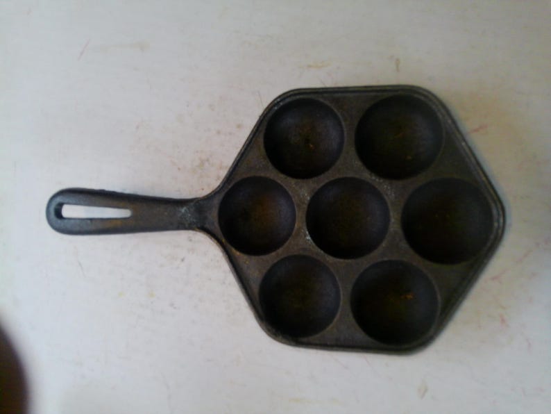 Może przedstawiać: Żeliwna patelnia w kolorze ciemnoszarym z siedmioma okrągłymi wgłębieniami do gotowania. Patelnia ma długi uchwyt z otworem do zawieszania. Powierzchnia patelni sugeruje, że jest dobrze wypalona i gotowa do użycia.
