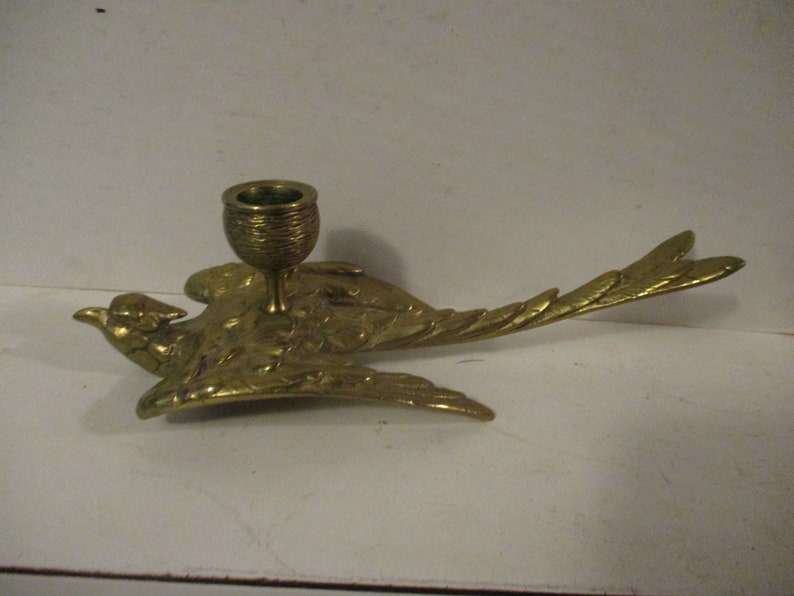 K&ouml;nnte beinhalten: Ein Messing-Kerzenhalter in Form eines Vogels im Flug. Der Vogel hat ausgebreitete Fl&uuml;gel und ein detailliertes Federdesign. Der Kerzenhalter ist eine kleine Tasse, geeignet f&uuml;r eine einzelne Kerze. Das Design ist vintage und dekorativ.