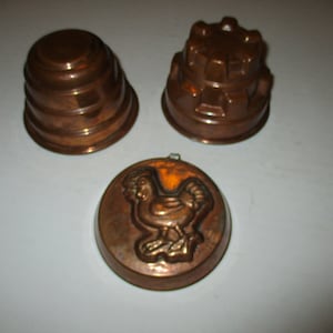 May include: Three vintage copper cake molds. One is a round mold with a raised design of a chicken. Another is a round mold with a tiered design. The third is a round mold with a castle-like design.