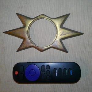 May include: A brass-colored, star-shaped metal object with a circular center cutout, positioned above a black remote control featuring a prominent purple circular navigation button and a red power button, providing a clear sense of scale.
