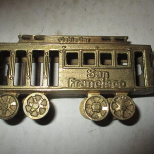 May include: A gold-colored metal cable car souvenir with the words "Cable Car" and "San Francisco" embossed on the side. The trolley has four wheels and a detailed design.