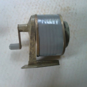 May include: A vintage metal pencil sharpener with a gray plastic crank and a metal base. The sharpener has a metal handle and a gray plastic blade.