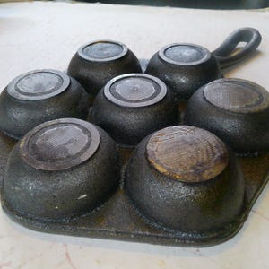 Może przedstawiać: Żeliwna forma do muffin&oacute;w z sześcioma zaokrąglonymi wgłębieniami. Patelnia jest ciemnoszara z teksturowaną powierzchnią i uchwytem. Wgłębienia mają okrągłe, teksturowane pokrywki. Idealna do pieczenia babeczek lub małych ciast.