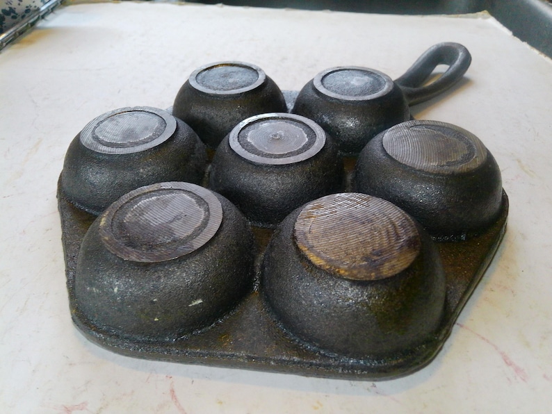 Może przedstawiać: Żeliwna forma do muffin&oacute;w z sześcioma zaokrąglonymi wgłębieniami. Patelnia jest ciemnoszara z teksturowaną powierzchnią i uchwytem. Wgłębienia mają okrągłe, teksturowane pokrywki. Idealna do pieczenia babeczek lub małych ciast.